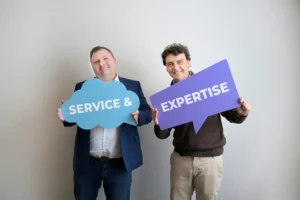 Two men in smart-casual attire smile while holding signs: a blue cloud reading 'SERVICE &' and a purple sign reading 'EXPERTISE'.