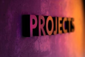 Close-up of three-dimensional letters spelling PROJECTS on a textured wall, lit with purple and orange light.