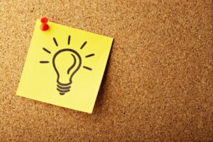 Yellow sticky note with a drawn light bulb pinned to a corkboard with a red pushpin, symbolizing an idea