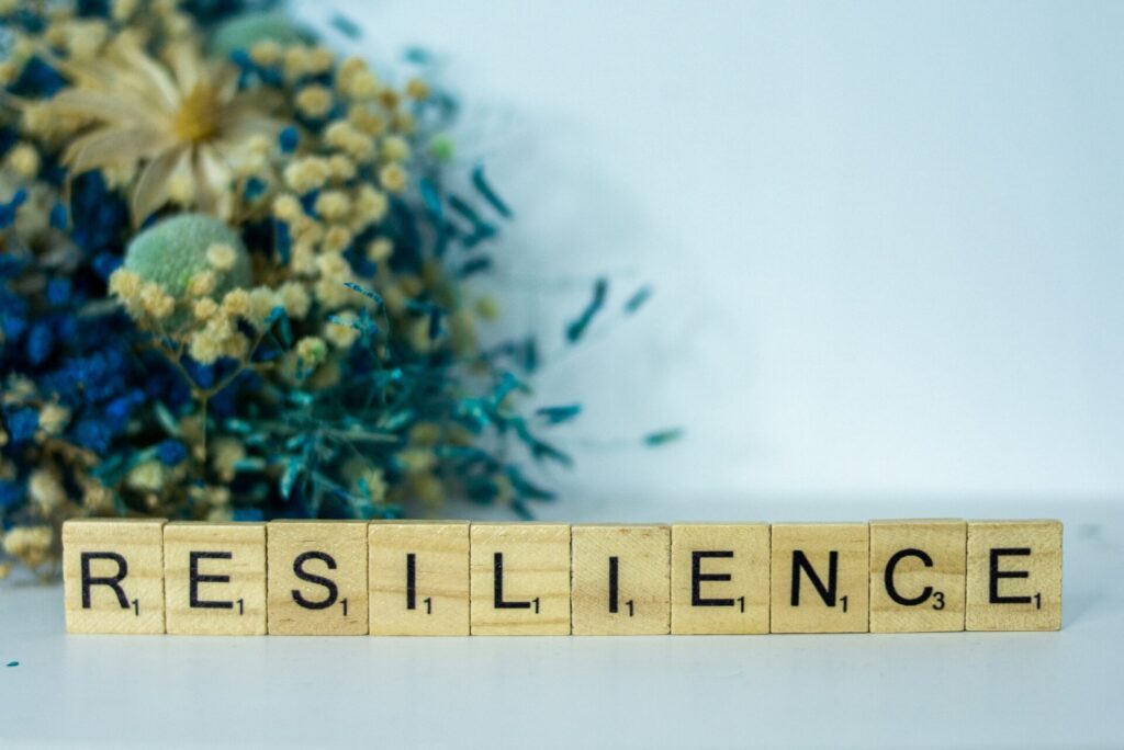 Row of Scrabble tiles spelling RESILIENCE on a light surface with a blurred bouquet in the background.