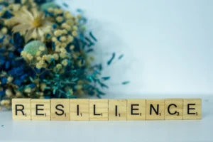 Row of Scrabble tiles spelling RESILIENCE on a light surface with a blurred bouquet in the background.