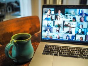 Laptop on a wooden desk displaying a multi-person video conference grid, with a teal ceramic mug in the foreground.