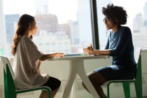 Two women sit across from each other at a bright office desk, chatting by a large window with a city skyline view.