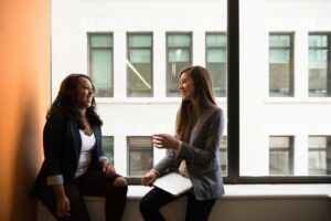 Two businesswomen sit by a large window, enjoying a friendly conversation; one holds a closed laptop on her lap.