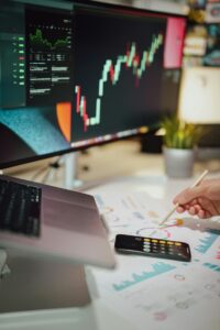 Trading desk with a monitor displaying a stock candlestick chart; a hand is drawing on a printed financial chart with a pencil, calculator nearby.