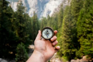 Hand holding a compass up in a forest, with blurred green trees in the background.