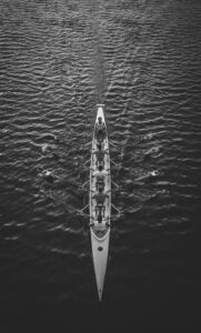 Aerial, black-and-white view of an eight-person rowing shell with rowers and oars on dark water.