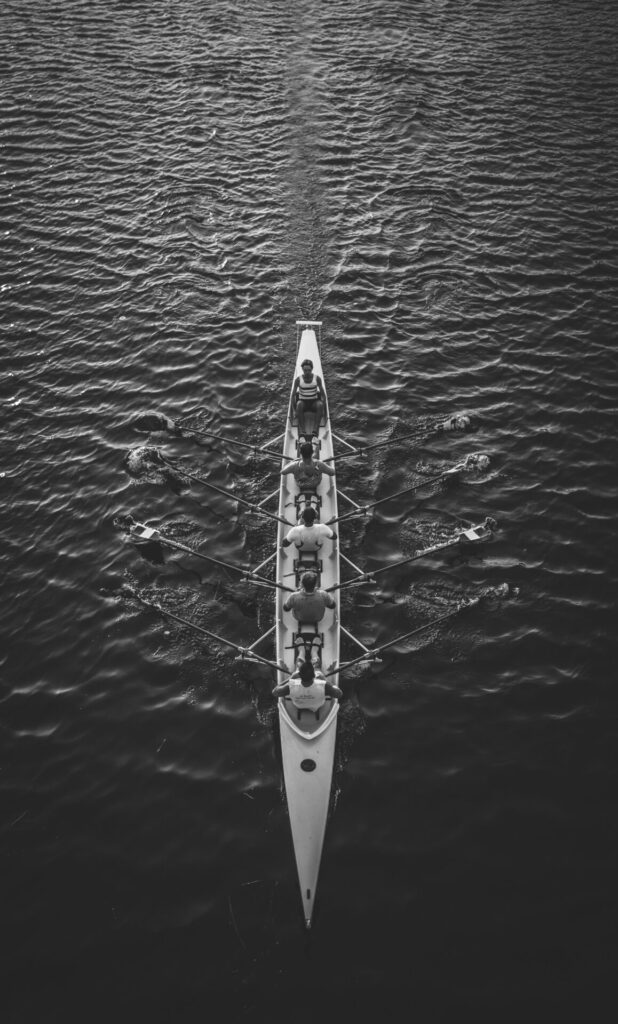Aerial, black-and-white view of an eight-person rowing shell with rowers and oars on dark water.