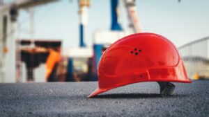 Red construction hard hat resting on a rough surface with blurred industrial structures in the background, suggesting a construction site safety scene.